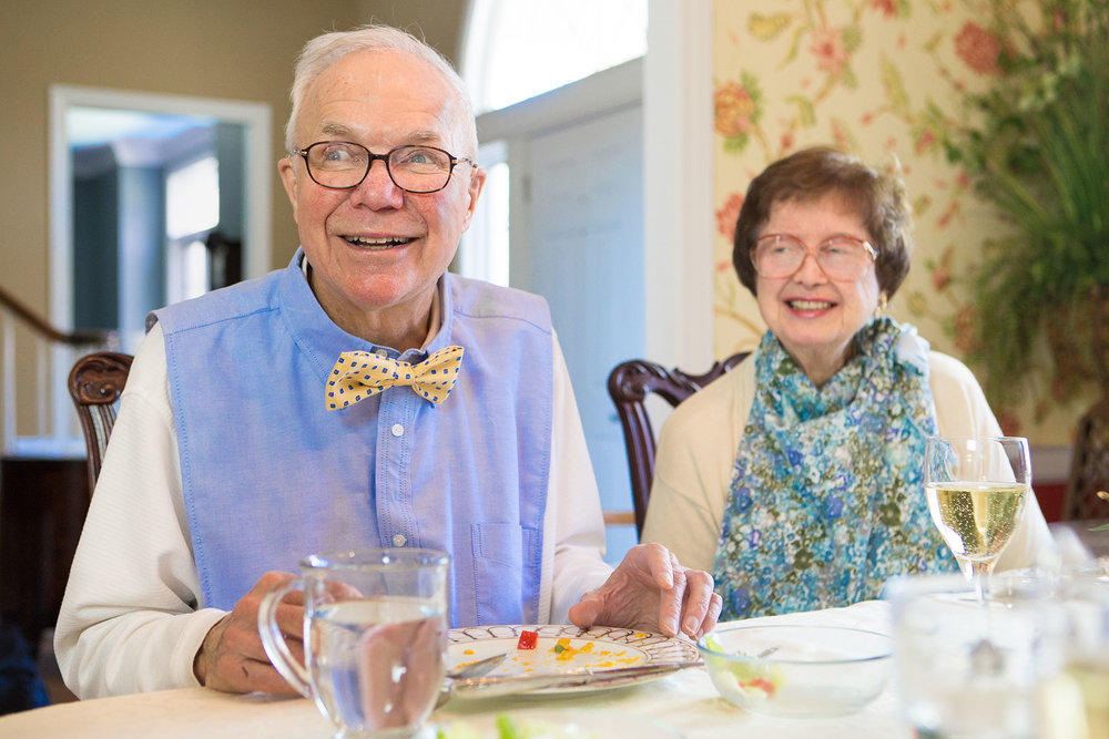 Older man sitting eating food with a white shirt covered by a light blue button up Dignity Bib with a yellow bow tie with red polka dots. He is sitting next to an older woman smiling with a cream shirt and a blue and green floral Dignity Bib that resembles a scarf around her neck.